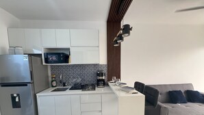 Una cocina moderna con armarios blancos, un refrigerador de acero inoxidable, un microondas integrado y una cafetera. En la sala de estar, hay un sofá gris con cojines azules.