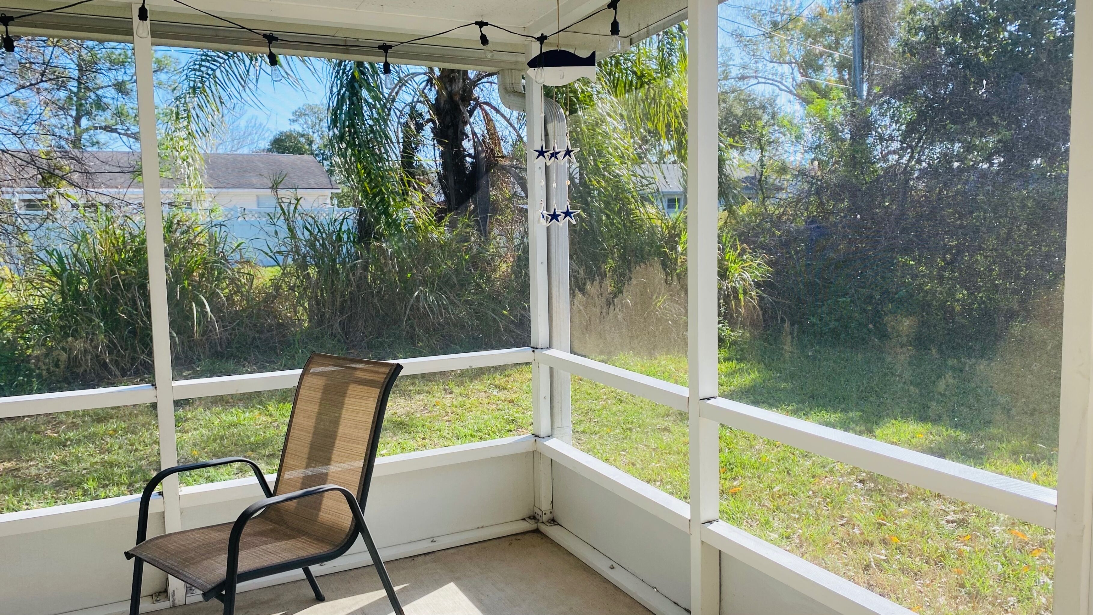 Ein überdachter Patio-Bereich mit einem einzelnen Sessel, großen Fenstern und Blick auf Grünflächen sowie ein Haus.