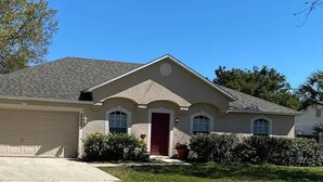 A single-story house with a garage, a red front door, and a small front yard with bushes and a tree. - Near Beach-3br-sleeps6-patio-garden-parking-ac (Edgewater)