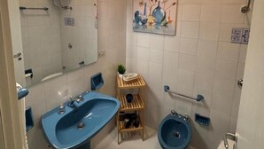A bathroom with a blue sink, a bidet, and a toilet. There is a mirror, a shelf with toiletries, and a painting on the wall. - Casa Maruzza - in the Centre of Cefalù (Cefalù)