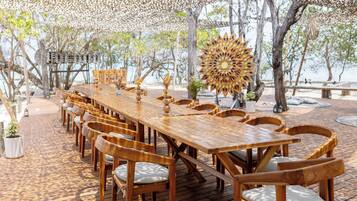 A wooden deck with a long table and chairs, surrounded by trees and a decorative sun-like structure.
