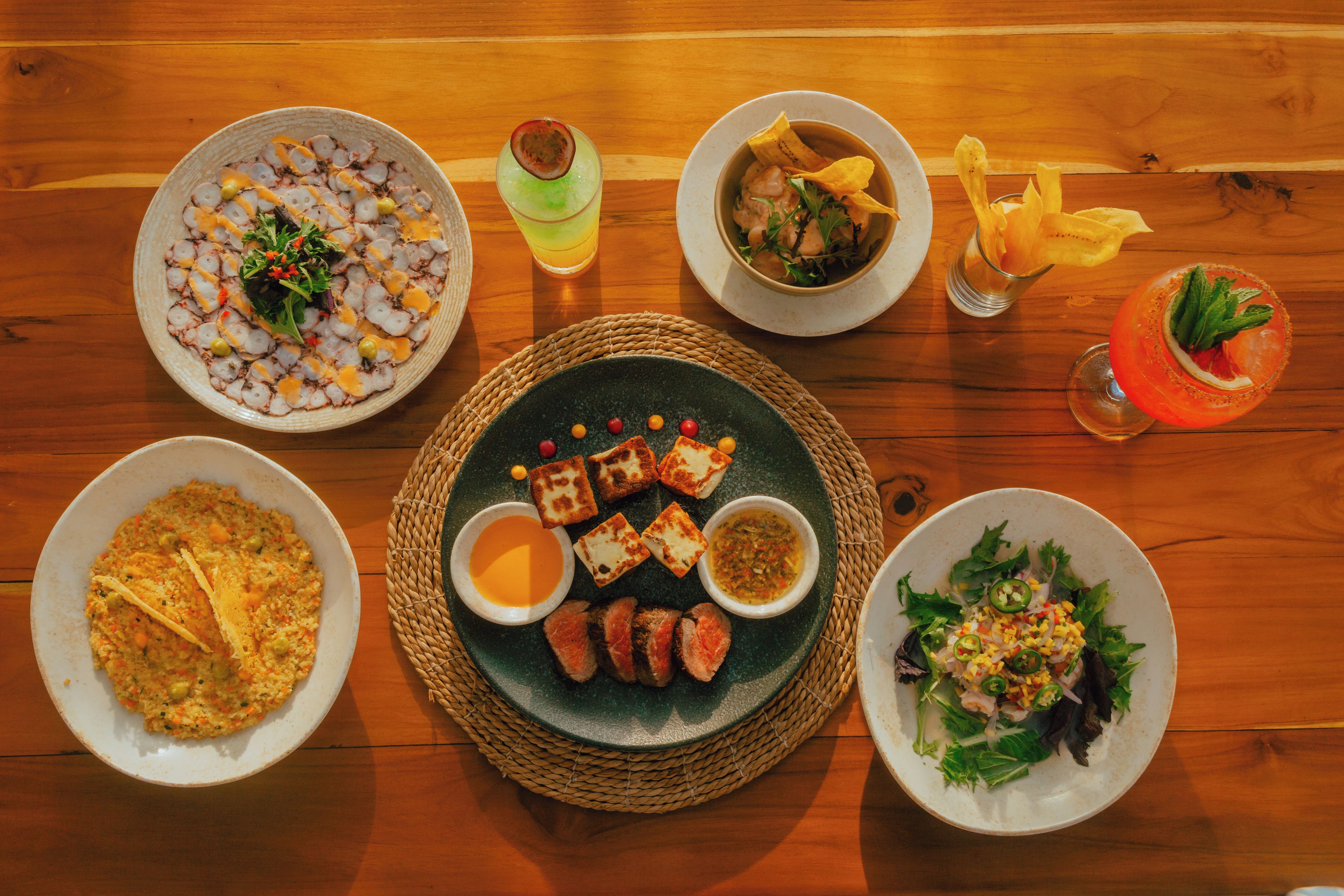 A variety of dishes on a wooden table, including a plate with octopus, a bowl with a green drink, and a plate with a salad.
