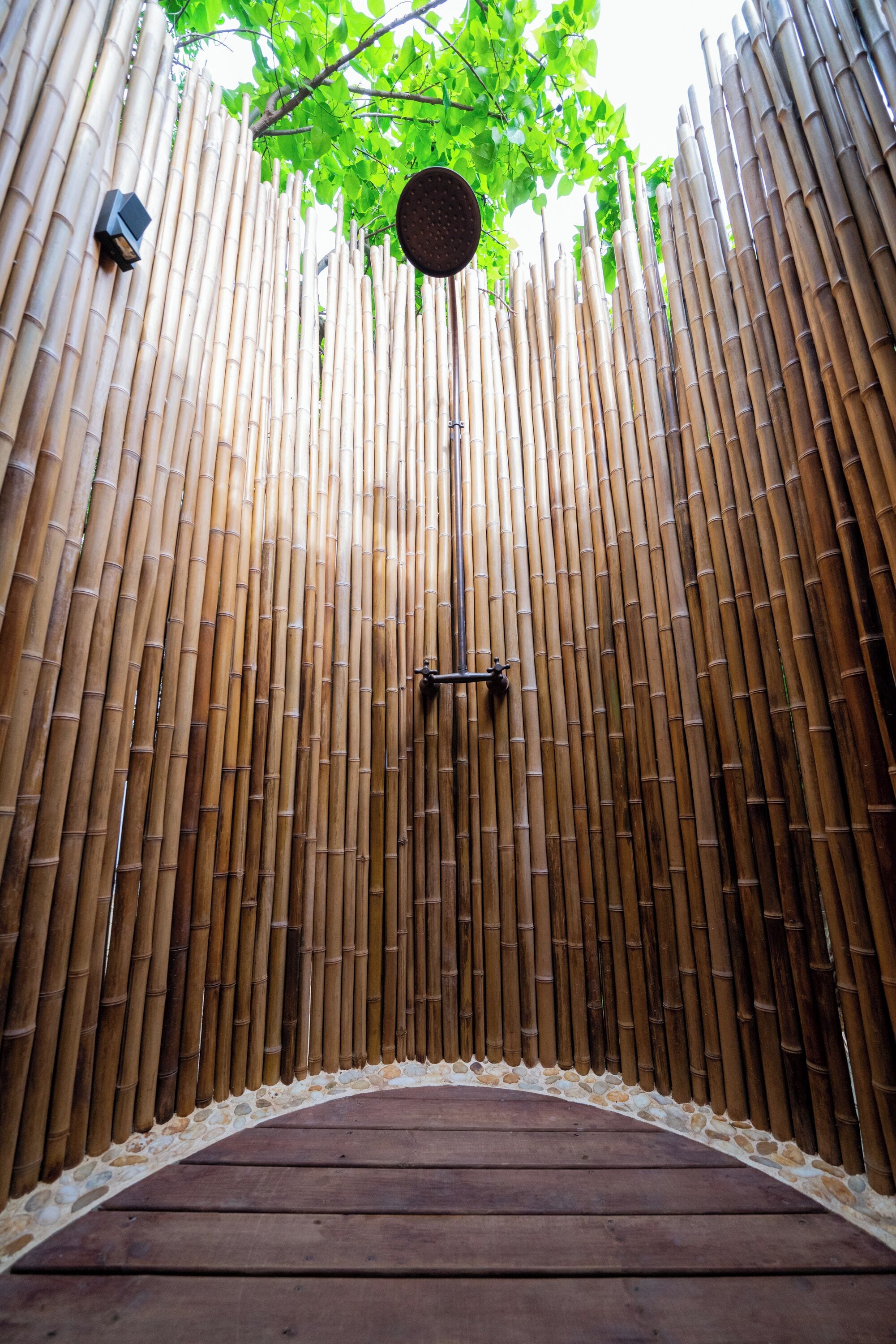 A bamboo wall with a circular opening, a wooden deck, and a hanging showerhead.