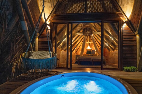 Une pièce chaleureuse, avec un toit en bois, un jacuzzi, un fauteuil en osier et un lit doté d’une table de chevet.