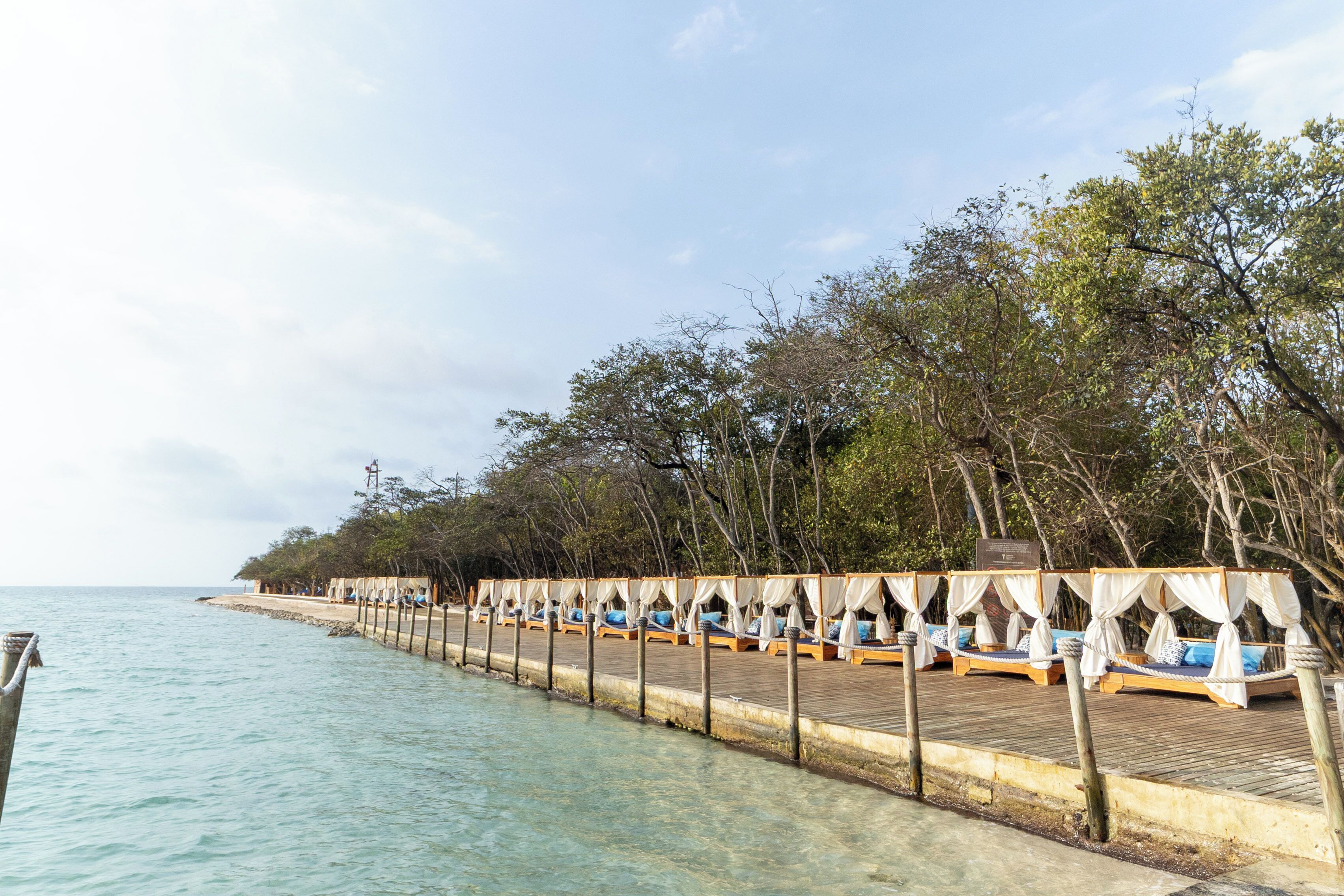 A wooden pier with rows of wooden beds with white canopies, overlooking clear turquoise water.