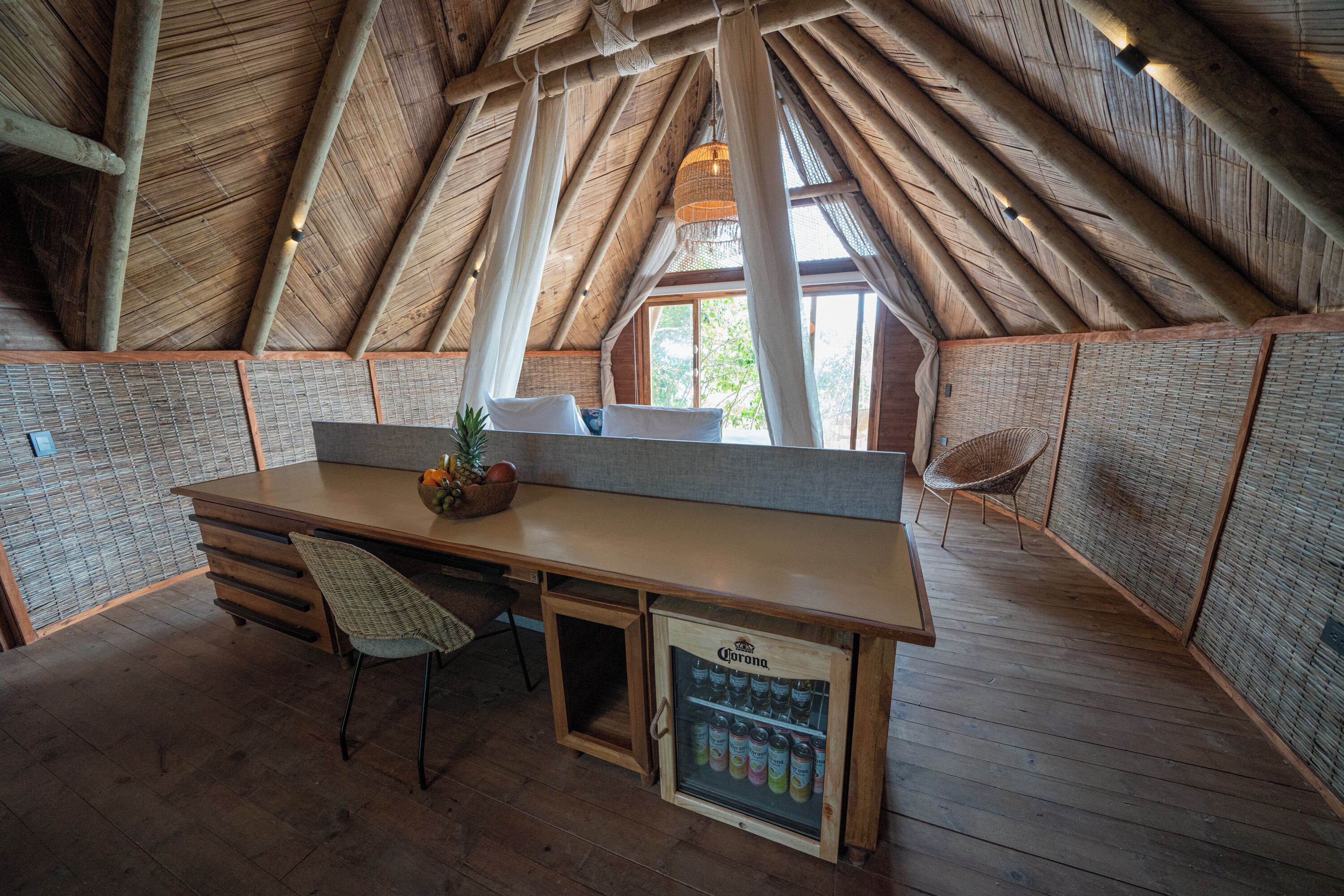 A rustic wooden interior with a high ceiling, a wooden desk, a chair, a small refrigerator, and a window with a view of trees.
