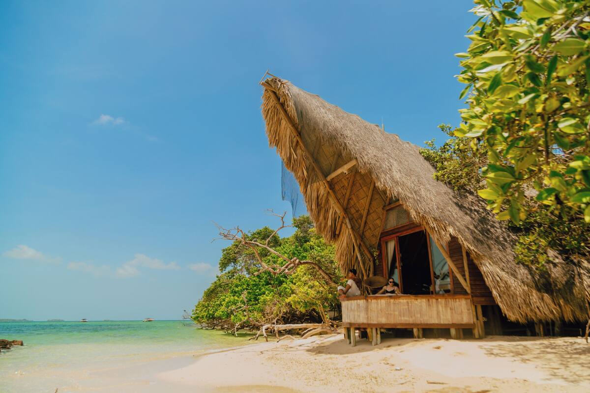 Eine strohgedeckte Hütte an einem sandigen Strand mit klarem, blauem Wasser und Grünpflanzen im Hintergrund.
