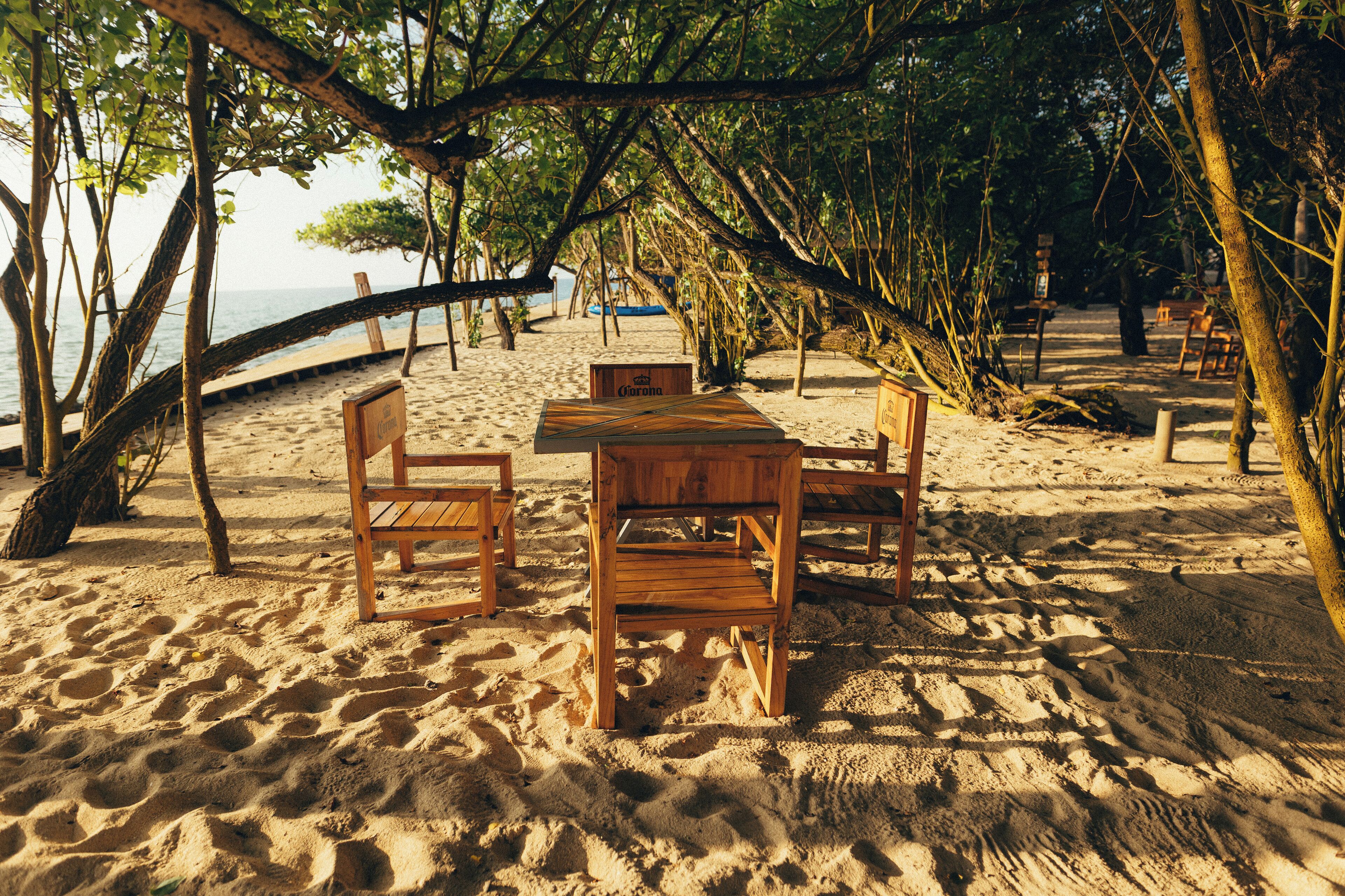 A wooden table and chairs set up on a sandy beach with a view of the ocean and trees in the background.