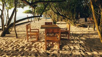 Un conjunto de mesa y sillas de madera sobre una playa arenosa, con vista al océano y árboles en el fondo.