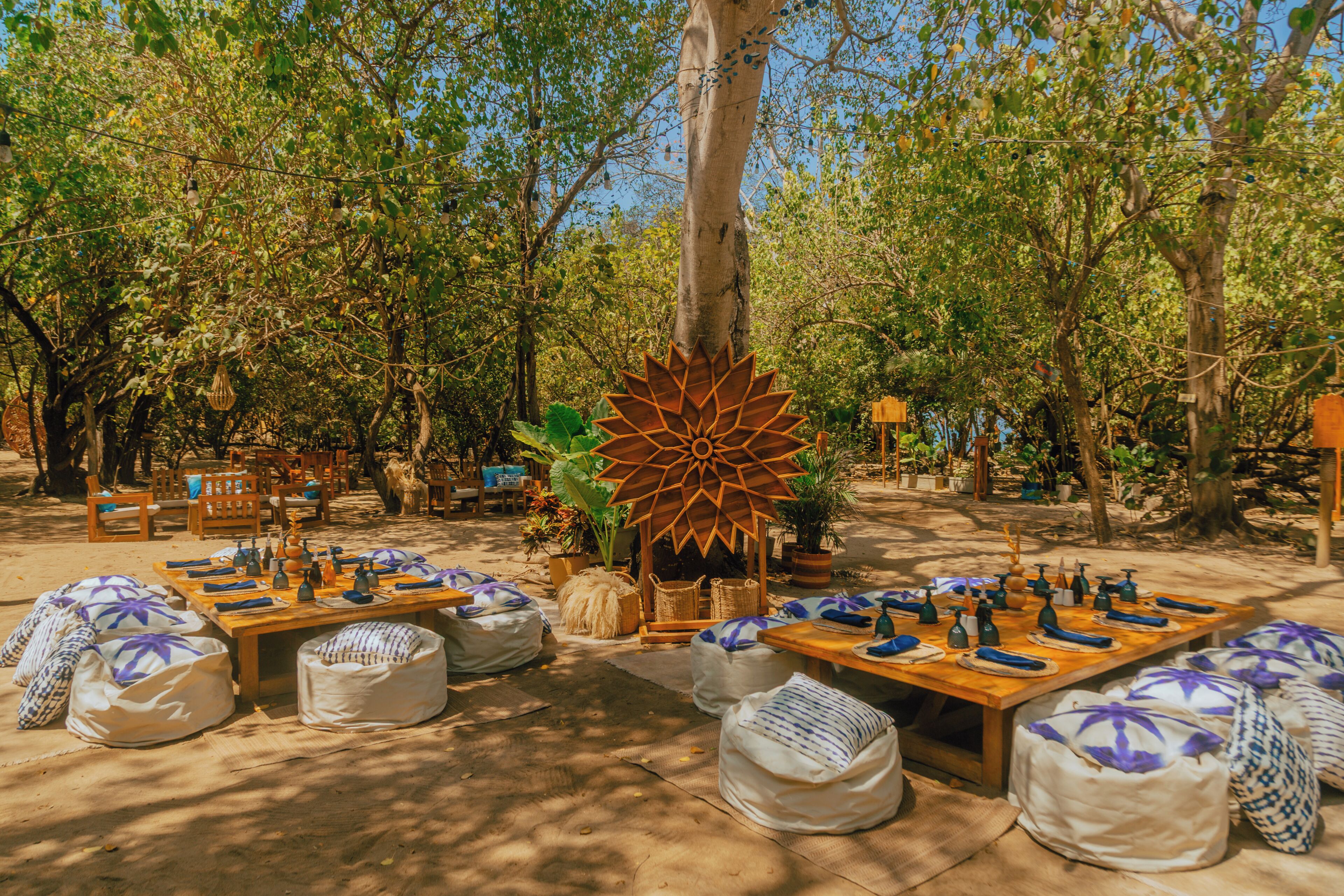 An outdoor dining area with wooden tables and benches, surrounded by trees and potted plants.