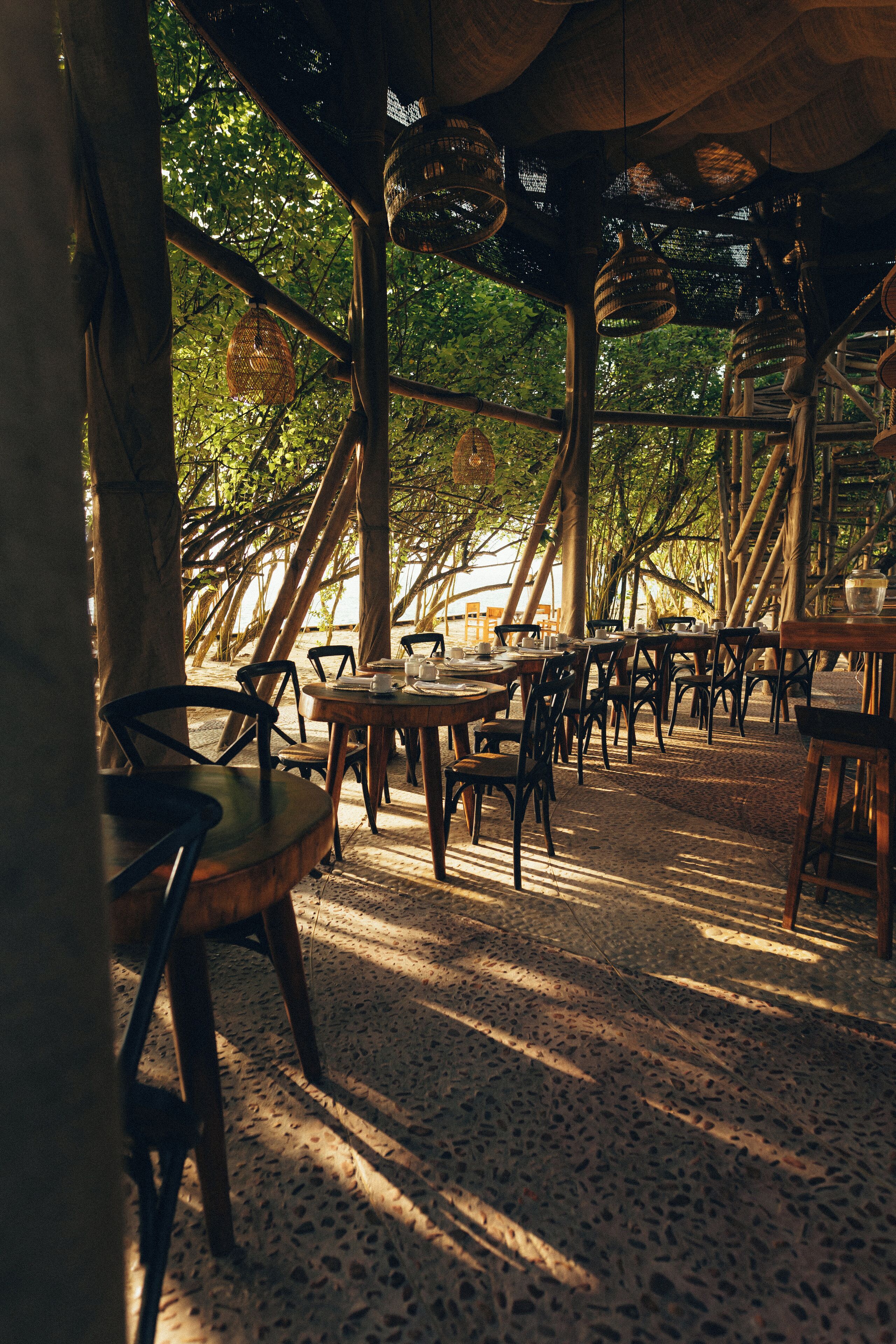 Un área de comedor al aire libre con mesas y sillas de madera, rodeada de árboles y una estructura de techo de paja.