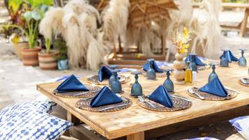 A wooden dining table set outdoors with blue napkins, a decorative centerpiece, and potted plants in the background.
