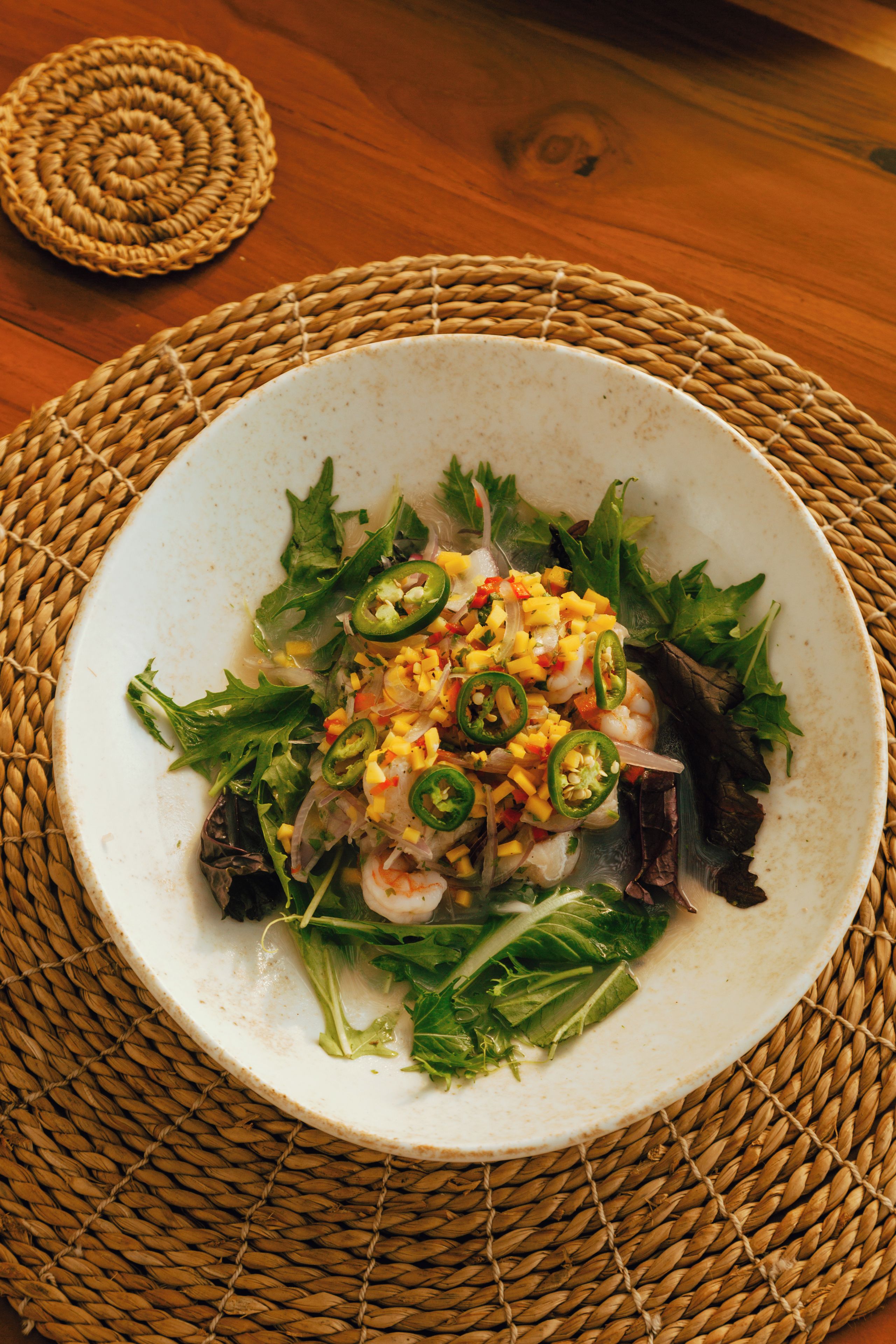 Une assiette de nourriture contenant des crevettes, de la mangue et des légumes verts, le tout posé sur un set de table en fibres tressées.