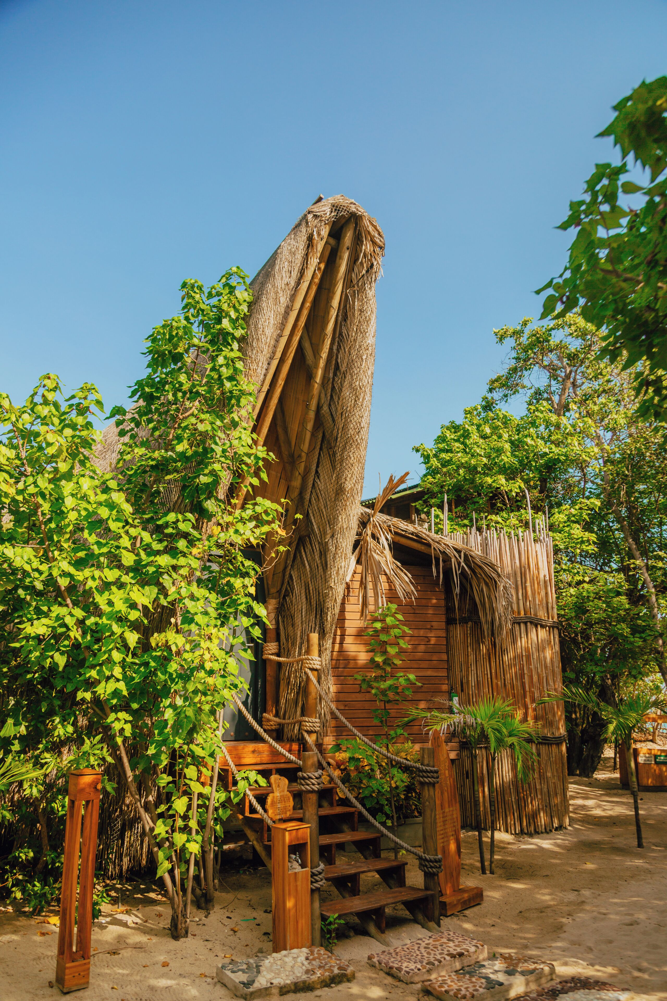 A rustic wooden cabin with a thatched roof, surrounded by lush greenery and a wooden staircase leading to the entrance.