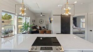 A modern kitchen with a central island, pendant lights, and a view to a living area with a sofa and a clock on the wall.