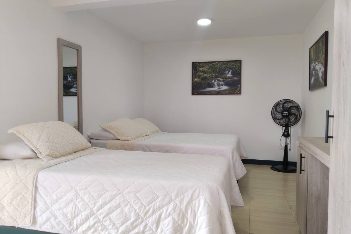 Een slaapkamer met twee bedden, een ventilator en een schilderij aan de muur.