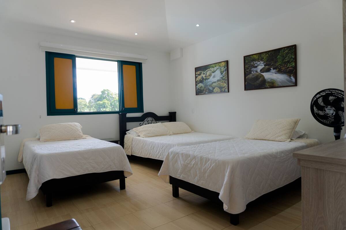 Een kamer met twee eenpersoonsbedden, een raam met gele omlijsting en twee wandventilatoren.