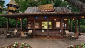 Un edificio de madera rústico con el rótulo "Woody grill and pool bar", con mesas al aire libre y un patio cubierto.