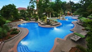 Una gran piscina curvada con sillones de baño y sombrillas, rodeada de árboles y vegetación.