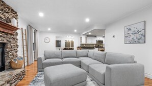 Un salon moderne avec un canapé d’angle, une cheminée en pierre et une cuisine en arrière-plan.