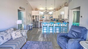 A modern living room with a sofa and a blue armchair, a kitchen with white cabinets and a brick backsplash, and a dining area with blue chairs.