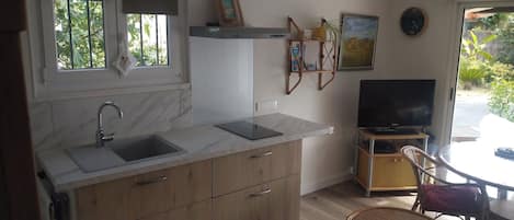 Una cocina con un lavamanos, encimera y armarios de madera. Hay un televisor sobre un mueble y una silla en la esquina.