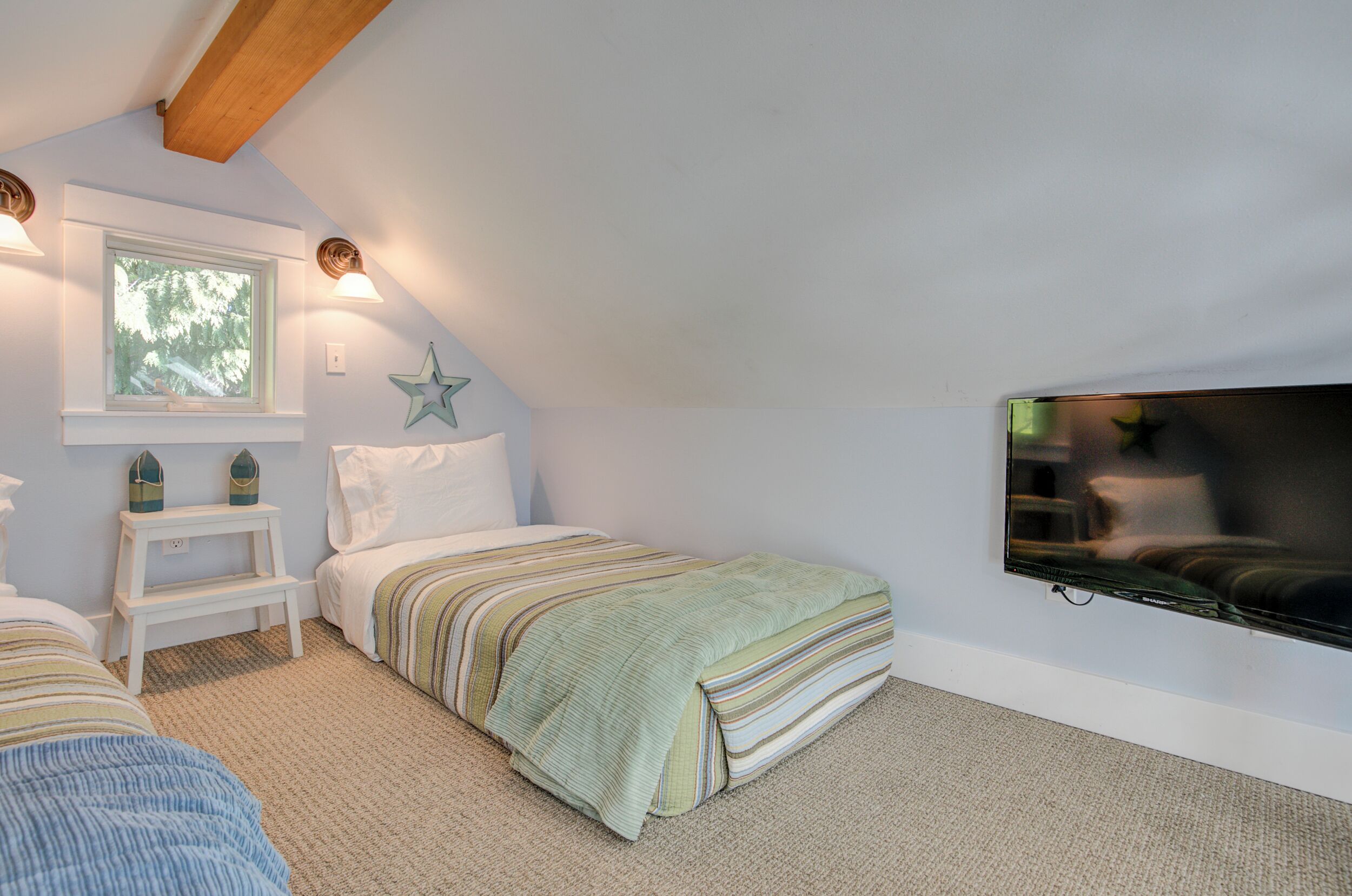 A bedroom with a sloped ceiling, a bed with striped bedding, a small white nightstand, a window with a view of trees, and a wall-mounted TV.