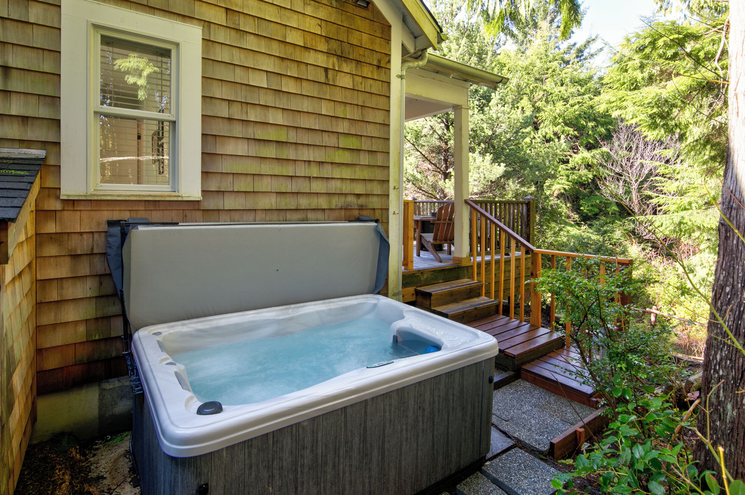 A hot tub outside a wooden house with a porch and steps leading to a deck.