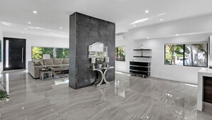 A spacious living room with a modern design, featuring a large central pillar, a sofa set, a console table with decorative items, and a large window providing a view of greenery.