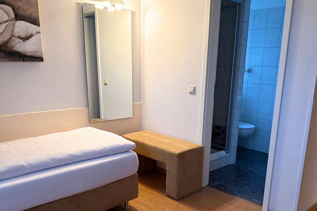 A bedroom with a bed, a bench, a mirror, and a bathroom visible through an open door.