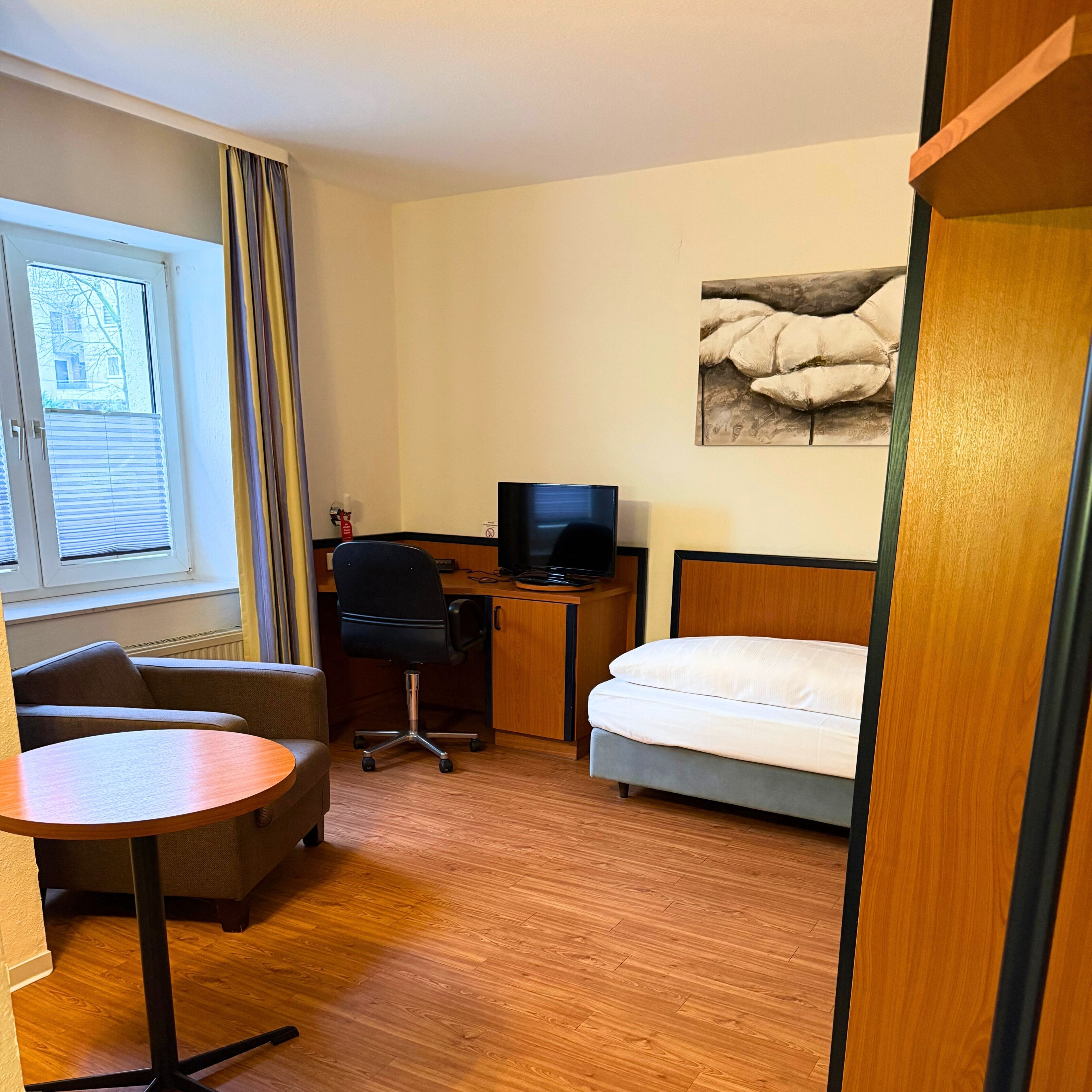 Une chambre d’hôtel comprenant un lit, un bureau avec un ordinateur, une chaise, une petite table et une fenêtre avec des stores.