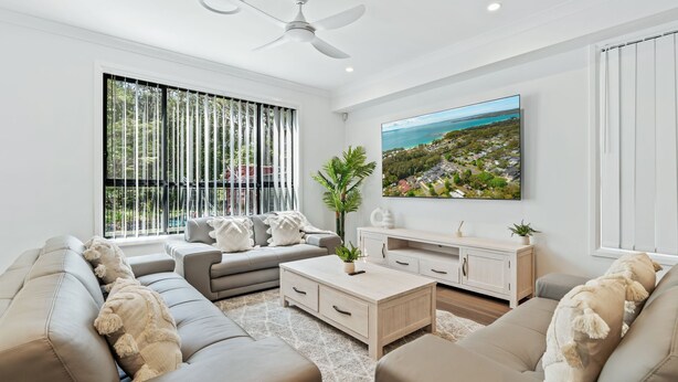 A modern living room with a large window, a ceiling fan, a flat-screen TV, and a light-colored sofa set. - La Casa by the Sea, Jervis Bay (Huskisson)
