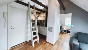 Un appartement de style « loft » avec des poutres apparentes, un coin cuisine, un espace salon avec un canapé et un escalier menant à une mezzanine.