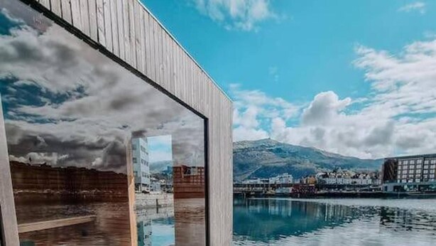 Un bâtiment moderne doté d’une terrasse en bois qui donne sur une étendue d’eau, avec une montagne en arrière-plan.