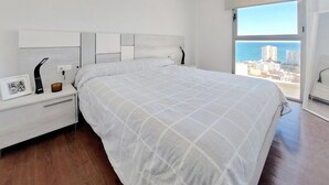 A bedroom with a large bed, a nightstand with a lamp, a framed photo, and a view of the city through a large window. - Beautiful home in Cullera (Cullera)