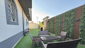 Un patio con mobiliario de mimbre y césped artificial, rodeado por un muro con listones de madera verticales y un seto.