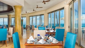 Un comedor amplio con techos altos, ventanas grandes que ofrecen vistas al océano y varios ventiladores de techo.