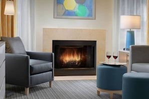 Een moderne woonkamer met een open haard, twee fauteuils, een bijzettafeltje met glazen en een schilderij aan de muur.