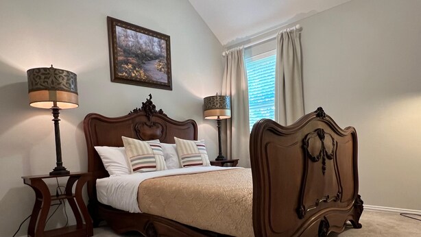 A bedroom with a large bed, two bedside tables, a nightstand, a painting on the wall, and a window with curtains. - Private Ground-Floor 2BR Suite in Gated Community - Near the Woodlands (Conroe)
