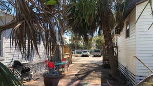 A narrow pathway leading to a patio area with a grill and seating, surrounded by palm trees and a white building. - Cozy Cottage, 1 bedroom sleeps 2 (Bradenton Beach)