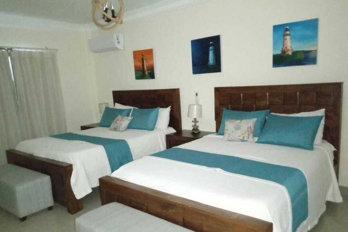 A hotel room with two beds, each with a wooden headboard and white bedding. There are two bedside tables with lamps, and a window with curtains.