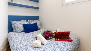 Un lit bien fait, recouvert d’une couvre-lit à motifs bleus et blancs, avec un oreiller bleu et un plateau rouge contenant des chocolats et des fleurs.