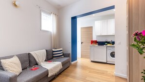 Un salon moderne avec un canapé gris, une couverture blanche et un coussin à rayures. Une fenêtre avec des rideaux blancs est visible, et on aperçoit au fond un coin cuisine avec un lave-linge.
