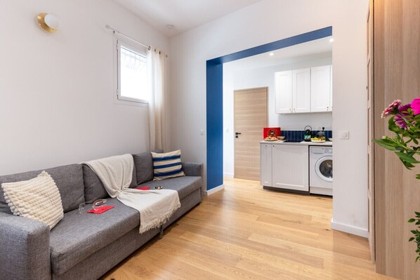 Un salón moderno con un sofá gris, una manta blanca y un cojín a rayas. Se ve una ventana con cortinas blancas y, al fondo, una zona de cocina con lavadora.