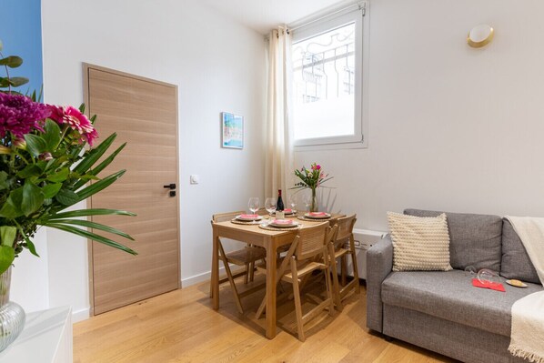 Una sala de estar acogedora con un sofá gris, una mesa de comedor de madera para dos personas y una ventana con cortinas.
