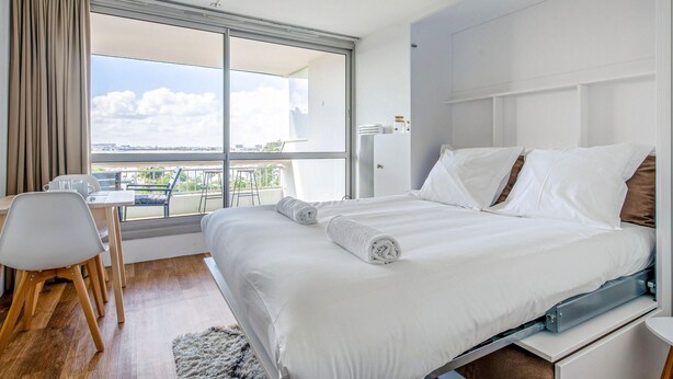 A modern hotel room with a large bed, a desk with a chair, a view of the city through a large window, and a balcony. - Cozy apartment in La Rochelle with WiFi (La Rochelle)