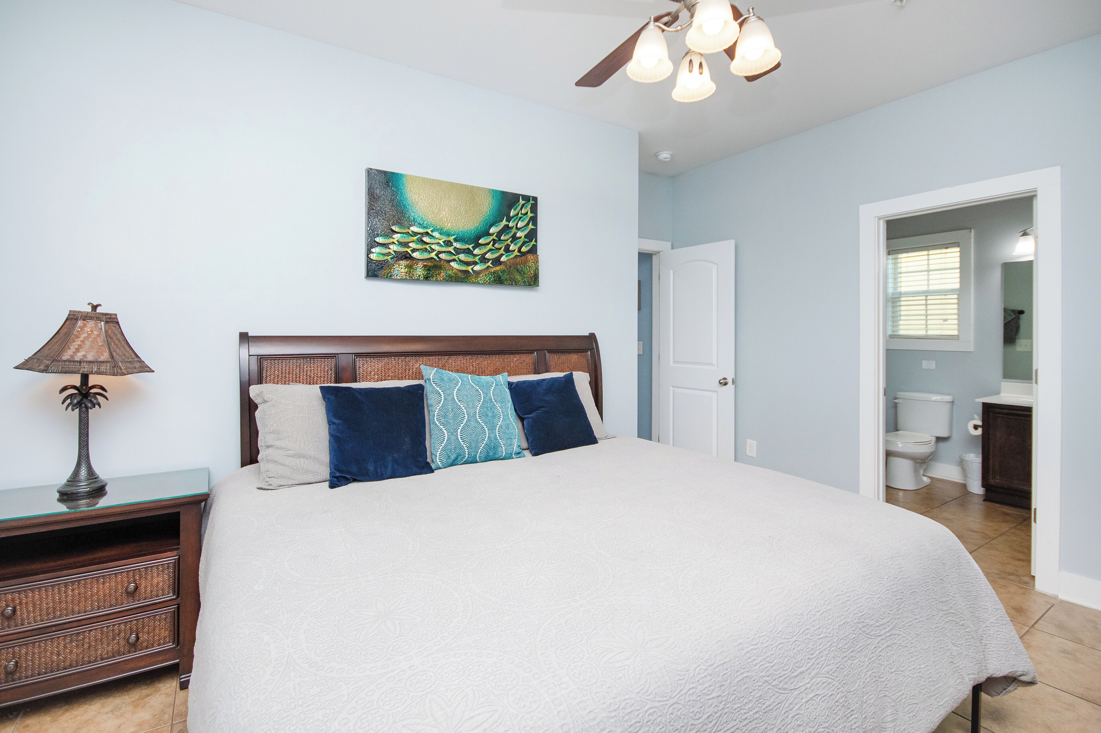 Une chambre à coucher avec un lit, une table de chevet, une lampe, un tableau, et une salle de bain visible à travers une porte ouverte.
