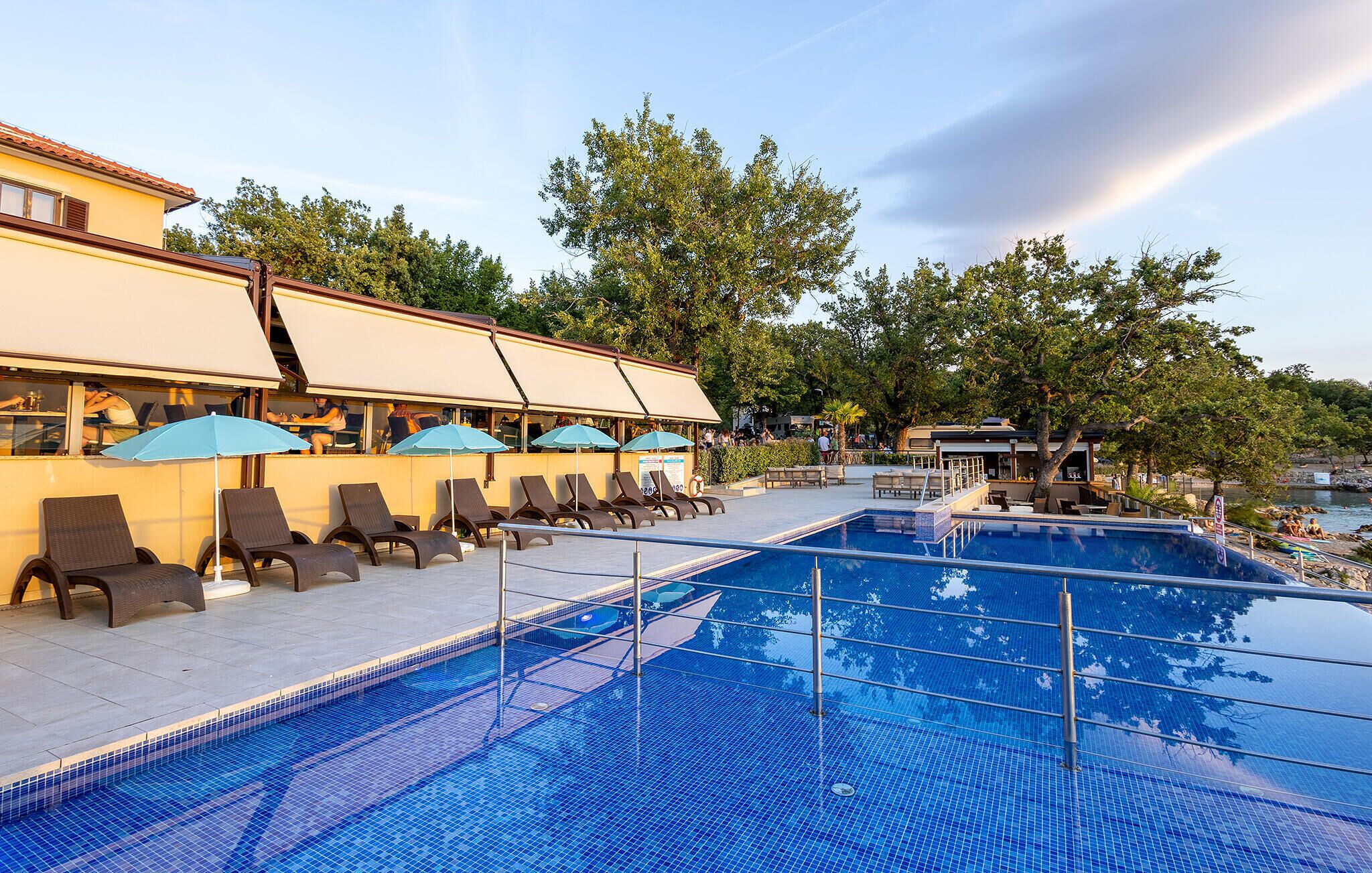 A swimming pool with lounge chairs and umbrellas, a restaurant with outdoor seating, and a view of the sea.