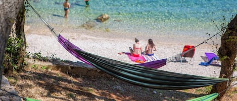 Una hamaca atada entre árboles en una playa con agua cristalina y gente al fondo.