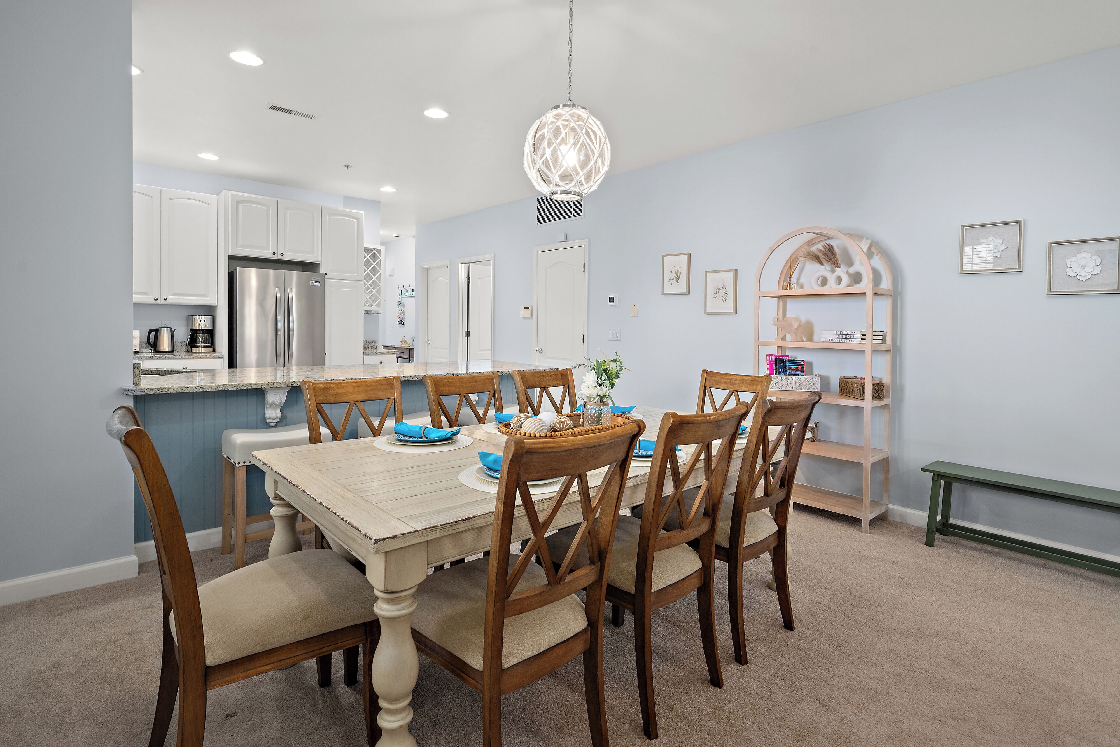 A dining area with a wooden table and chairs, a chandelier, and a kitchen in the background.